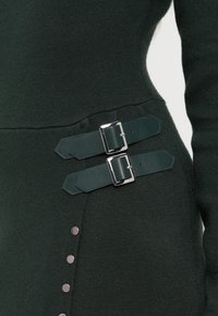 Tmavě zelené pletené šaty s dvěma odpovídajícími koženými popruhy se stříbrným kováním a akcenty knoflíků na boku.