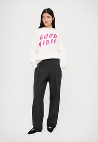 Maglione bianco a coste con "Good Vibes" in rosa neon, abbinato a pantaloni grigio scuro a vita alta e ballerine nere a punta.