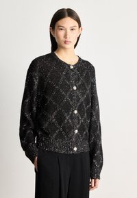Black cardigan with diamond pattern in metallic thread, featuring round buttons and ribbed hem. Soft texture with long sleeves.