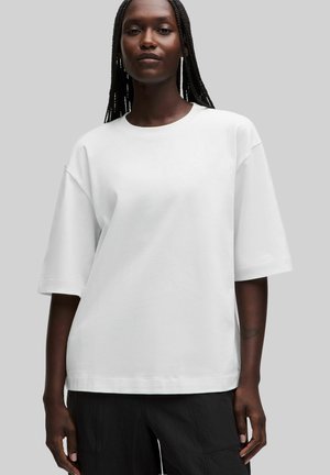 Dunkelhäutige Person mit langen geflochtenen Haaren trägt ein schlichtes weißes übergroßes T-Shirt und schwarze Hosen vor einem grauen Hintergrund.