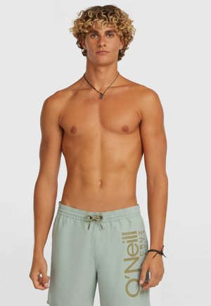 Junger Mann mit lockigem blondem Haar trägt hellgrüne Badeshorts mit O'Neill-Logo, Halskette und Armband, steht vor einem schlichten Hintergrund.