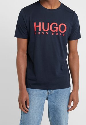 Mężczyzna noszący granatowy T-shirt z dużym czerwonym napisem „HUGO” i mniejszym „HUGO BOSS”, w połączeniu z jasnoniebieskimi jeansami.