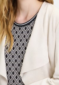 Kobieta w beżowej kurtce na zamek, nosząca czarno-białą bluzkę w geometryczny wzór, z długimi blond włosami widocznymi po lewej stronie.