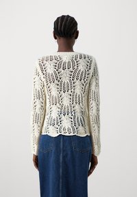 Pletený svetr v krémové barvě s volným krajkovým vzorem. Má kulatý výstřih, dlouhé rukávy a lem s vlnitým okrajem, stylizovaný přes džínsovou sukni.