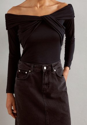 Vrouw draagt een zwarte off-shoulder top met lange mouwen en gedraaide details, gecombineerd met een hooggesloten zwarte denim rok met zakken.