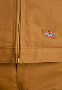 Brun arbejdsjakke med lynlås, lavet af kraftigt stof, synlig gul lynlås og et patch-logo på forsiden, der har et cirkulært design med tekst.