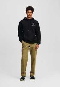 Schwarzer Hoodie mit Fronttasche, Kapuze mit Kordelzug und weißem Grafikdruck; kombiniert mit hellen Khaki-Hosen und schwarzen Sneakers mit gelben Akzenten.