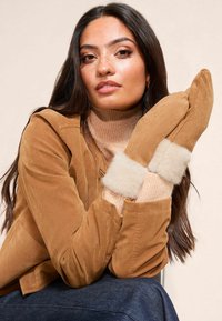 Pruun seemisnahast jakk, millel on soonikkoes beež kaelusega kõrgkaelusega kampsun, millele on lisatud beežad kinnased pehme karvase mansetiga. Nähtavad on ka tumedad teksapüksid.