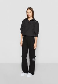 Black zip-front jacket with long sleeves and front pocket, paired with loose black cargo pants featuring a side patch pocket and white logo.