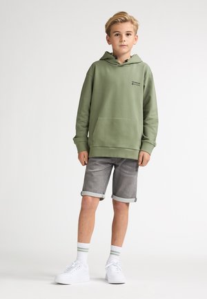 Hanorac verde măsliniu cu buzunar frontal și logo negru, combinat cu pantaloni scurți din denim gri cu tivuri rulotate. Adidași albi și șosete cu dungi.