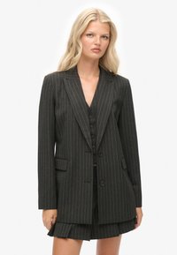Sort blazer med smalle striber, med klapkrave, skræddersyet pasform, forlommer og matchende vest. Båret med en plisseret nederdel.