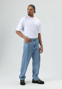 Witte katoenen T-shirt, ruimvallend, gecombineerd met lichtblauwe denim jeans met voorzakken. Model draagt zwarte schoenen met dikke zolen.