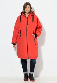 Parka rouge longue avec capuche, zippers et cordons de serrage noirs, deux poches avant et ourlet élastique. Portée sur un jean foncé et des bottes noires.