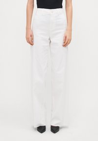 Pantalones de pierna ancha blancos hechos de algodón con cintura alta. Cuentan con un cierre de botón y un sutil detalle en las costuras laterales. Se llevan con zapatos negros de puntera.