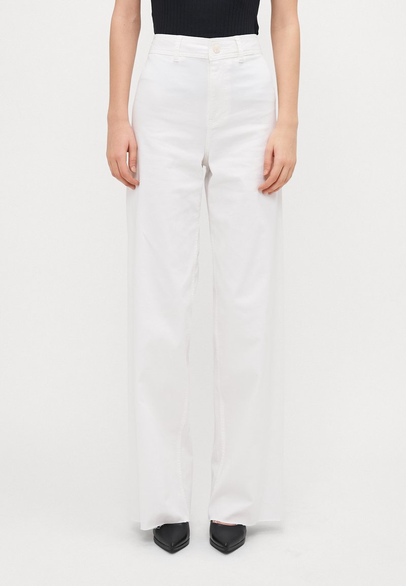 Pantalones de pierna ancha blancos hechos de algodón con cintura alta. Cuentan con un cierre de botón y un sutil detalle en las costuras laterales. Se llevan con zapatos negros de puntera.