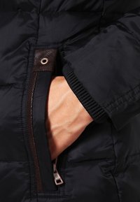 Svart isolerad jacka med en brun dragkedjeficka och ribbade ärmslut. Materialet har en slät, blank yta. Handen är i fickan.