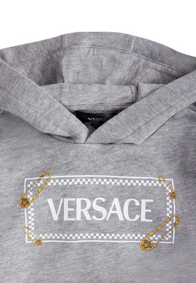 Un sweat à capuche gris en coton, affichant un grand logo blanc "VERSACE" avec une bordure à carreaux et des accents de épingle de sûreté dorée sur le devant.