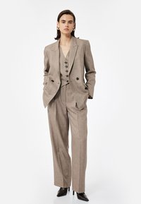 Beige op maat gemaakt pak met een doublé blazer, een drieknoop vest en wijde broekspijpen. Glad materiaal met een subtiele textuur.