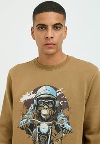 Brun sweatshirt med grafik af en monkey i en blå hjelm, der kører på motorcykel, med splatter-effekter i baggrunden.