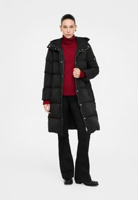 Cappotto imbottito nero con cappuccio, caratterizzato da un design trapuntato e chiusura con zip. Indossato sopra un dolcevita rosso e pantaloni neri.