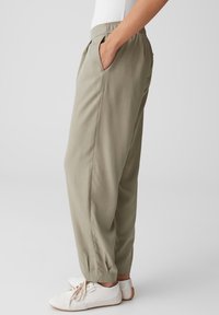 Person trägt hellbeige elastische Bundhose mit Seitentaschen und weiße Sneakers, von der Seite vor einem einfarbigen Hintergrund gezeigt.