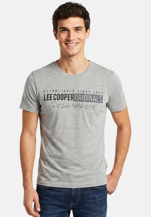 Jeune homme souriant portant un t-shirt gris « Lee Cooper Originals » et un jean bleu, debout devant un fond blanc uni.