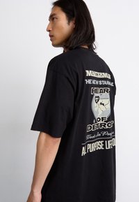 Černé oversized tričko vyrobené z bavlny, s grafickým textem a designem globu v tlumených barvách na zadní straně. Krátké rukávy, kulatý výstřih.