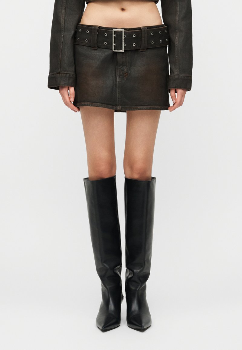 Femme portant une mini-jupe en cuir noir avec ceinture, une veste courte assortie et des bottes en cuir noir jusqu'aux genoux, debout devant un fond blanc.