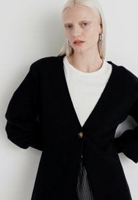 Black cardigan with a v-neck, oversized fit, and a single brass button. Material appears textured, worn over a white top.