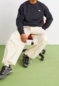 Černá mikina s logem, krémové volné kalhoty a černé sportovní boty se zelenými akcenty, sedící na otočné židli na zelené podlaze.