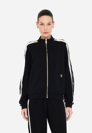 Donna con i capelli pettinati all'indietro, che indossa una giacca nera con zip e pantaloni abbinati con strisce bianche ai lati, su uno sfondo bianco uniforme.