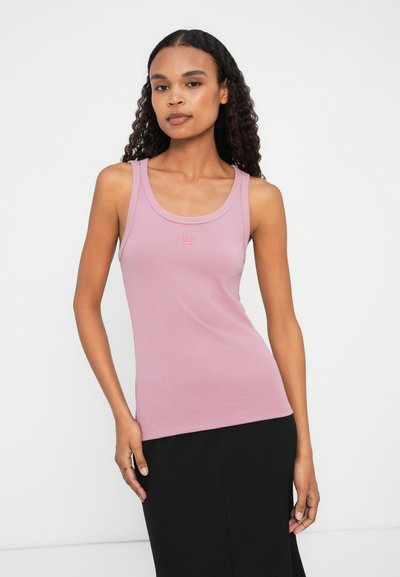 Débardeur rose clair en matériau doux, avec un décolleté en cœur et un petit logo au centre. Le modèle l'associe avec une jupe noire.