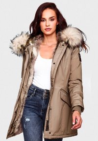 Beige parka met een nepbontcapuchon, rits- en drukknoopsluiting, zijzakken, geribbelde manchetten en een zachte voering. Dracht over een witte top en jeans.