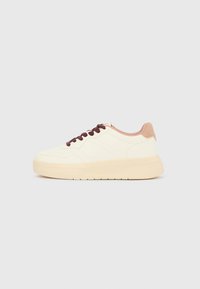 Baskets basses beiges avec semelle épaisse, bout perforé, col de talon rose et lacets brun foncé sur un fond gris clair uni.