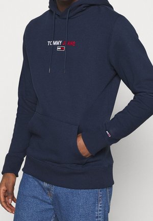 Marineblauwe hoodie met voorzak, "Tommy Jeans"-logo in wit en rood op de borst, klein vlaggenlogo op de linker manchet, gedragen met een blauwe spijkerbroek.