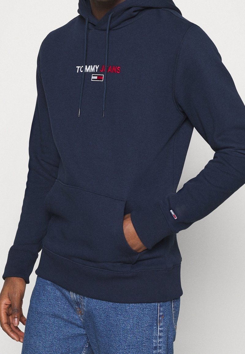 Navy blue hoodie with front pocket, "Tommy Jeans" logo in white and red on chest, small flag logo on left cuff, worn with blue jeans.