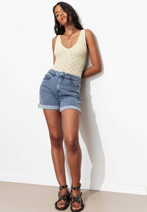Femme portant un haut en dentelle crème sans manches, un short en jean roulé, des sandales noires cloutées et un collier en or, debout contre un mur blanc.