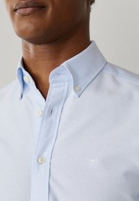 Primo piano di una persona che indossa una camicia azzurra con collo allacciato e un discreto logo ricamato sul petto.