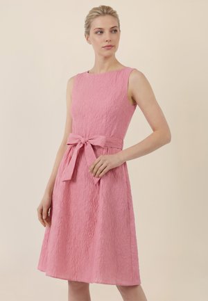 Kobieta w różowej, bezrękawnikowej sukience o fakturze z wiązaną kokardą w talii, stojąca na tle gładkiego beżowego tła, patrząca w swoją lewą stronę.