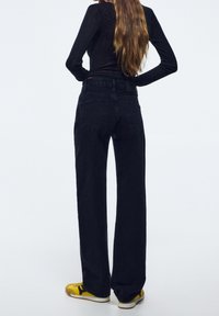 Jean noir taille haute à jambes larges avec deux poches arrière, texture lisse et détails de couture subtils, assorti à des baskets jaunes et blanches.