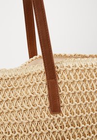 Vævet stråtaske i beige farve med teksturerede tråde, suppleret med brune læderhåndtag for holdbarhed og stil.