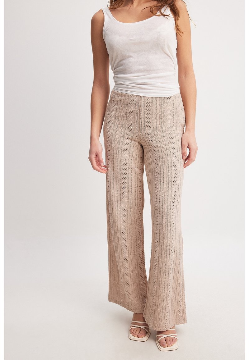 Pantalons beiges à large jambe en tissu tricoté texturé, présentant des rayures verticales et des découpes décoratives. Associés à un débardeur blanc ajusté.