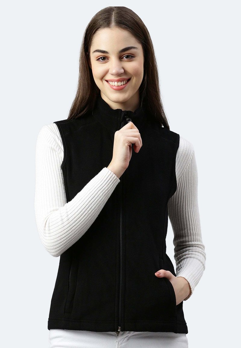 Schwarze Fleece-Weste mit hohem Kragen, frontalem Reißverschluss und Seitentaschen, über einem weißen gerippten Langarmoberteil getragen. Glatte Textur.