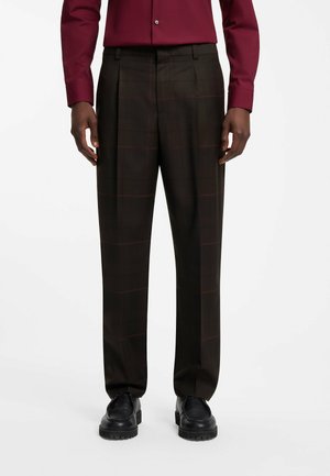 Hombre que lleva pantalones de cuadros marrón oscuro, zapatos de cuero negros y una camisa burdeos de manga larga con botones, de pie sobre un fondo blanco.