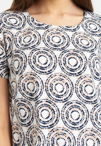 Lyhythihainen toppi, jossa on valkoinen tausta ja samansuuntaisia ympyröitä tummansinisessä, beigessä ja ruskeassa, valmistettu kevyestä kankaasta, jolla on sileä pinta.