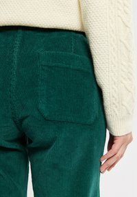 Un pantalon en velours côtelé vert dispose d'une poche arrière et d'une texture nervurée verticale, associé à un pull en tricot crème avec des détails en torsades.