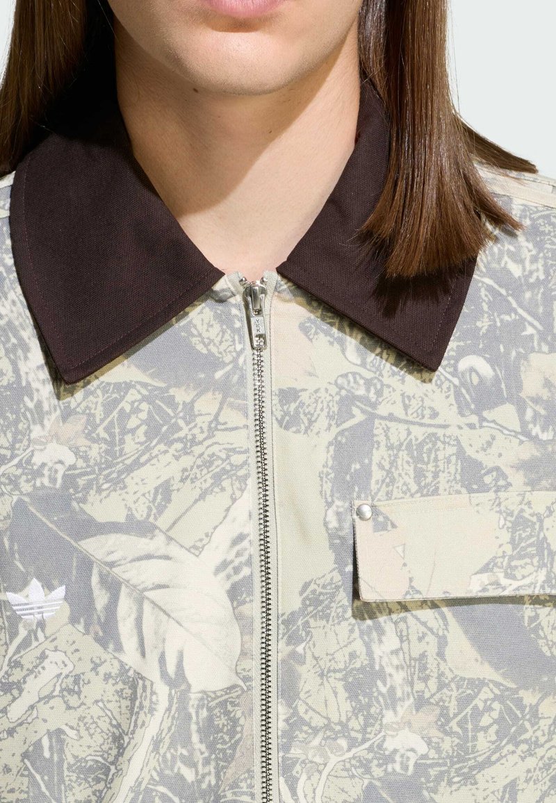 Reißverschluss-beige Jacke mit braunem Kragen, Tarnmuster und einer linken Brusttasche mit Druckknopf. Hergestellt aus strukturiertem Stoff.