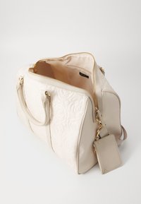 Sac à main matelassé beige avec doubles poignées, quincaillerie dorée et poches extérieures zippées. Intérieur doublé en tissu léger. Comprend une étiquette amovible.