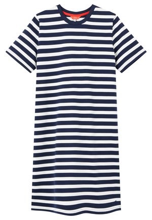 Frau trägt ein knieumspieltes, kurzärmeliges Kleid mit dunkelblauen und weißen horizontalen Streifen und weiße Turnschuhe, steht auf einem grauen Boden.