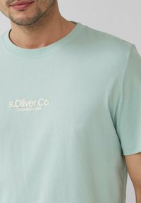 Jasnozielony bawełniany t-shirt z okrągłym dekoltem, z białym haftowanym napisem "s.Oliver Co. Established 1969" na piersi.